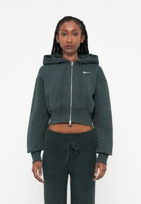 Tamnozelena skraćena hoodie sa rajsnicom, prednjim džepovima i malim bijelim logotipom. Uparen sa odgovarajućim tamnozelenim fluffy hlačama.
