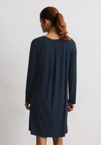 Donna con capelli rossi legati in una coda di cavallo, che indossa un vestito lungo, blu scuro, al ginocchio, in piedi di fronte a una parete bianca e texturizzata.