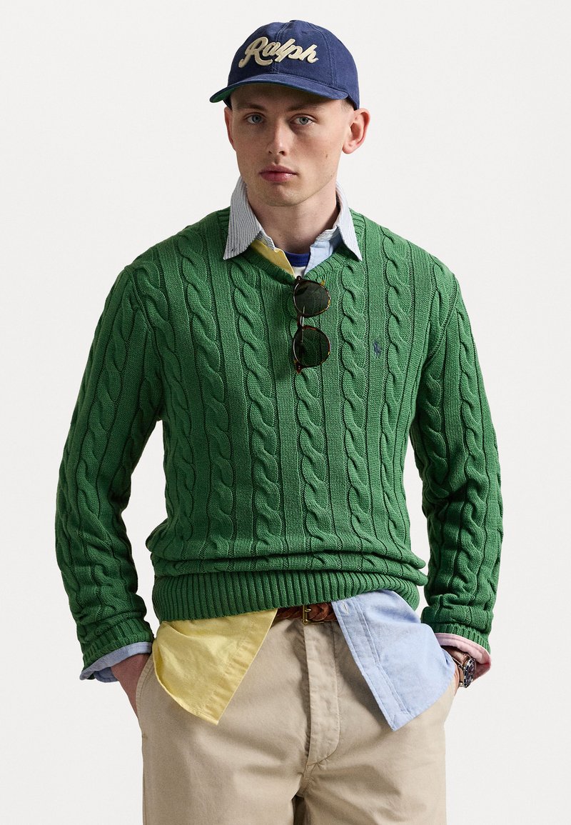 Green cable-knit sweater with a V-neck, layered over a blue and yellow collared shirt. Tan pants and dark sunglasses complete the outfit.