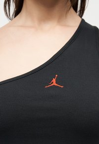 Zbliżenie osoby noszącej czarny prążkowany top na jedno ramię z małym czerwonym haftowanym logo Jumpman na piersi.