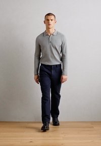 Světle šedá dlouhá polo košile, námořnické kalhoty, černé boty. Model se prochází s uvolněnýma rukama po stranách. Jednoduché, moderní prostředí.