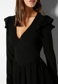 Schwarzes, strukturiertes Kleid mit tiefem V-Ausschnitt, langen Ärmeln mit Rüschendetails und einem ausgestellten Rock, der eine betonte Silhouette hervorhebt.