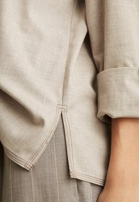 Lichtbeige shirtstof met een subtiele textuur; heeft een omgevouwen manchet en zichtbare naden aan de zoom met een gespleten ontwerp.
