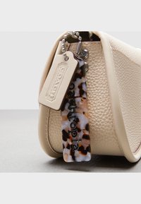 Beżowa skórzana torebka o fakturowanym wykończeniu, z zawieszką w ciemne i jasne kolory żółwia oraz metalowym breloczkiem.