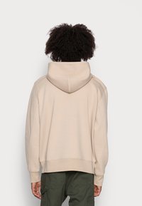 Sudadera con capucha beige con una capucha amplia, puños acanalados y un ajuste holgado; la tela parece suave y texturizada. Combinada con pantalones cargo verdes.