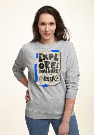 Šedá mikina s grafikou roztrženého papíru a černým textem "EXPLORUJ! JEŠTĚ VÍCE DOBRODRUŽSTVÍ", doplněná modrou páskou. Volný střih.