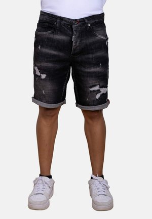 Persona que lleva puestos unos pantalones cortos vaqueros negros desgastados remangados en el bajo con zapatillas blancas sobre un fondo liso.