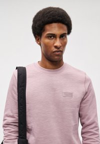 Gaiši rozā džemperis ar apaļu kakla izgriezumu, ribotu piedurkņu galiem un mazu uzlīmi uz krūšu. Gluda tekstūra ar smalku heathered rakstu.