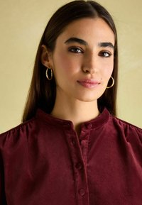 Bordeaux Samtbluse mit hohem Kragen, Knopfverschluss und gerafften Schultern. Das Modell trägt goldene Creolen vor einem hellen Hintergrund.