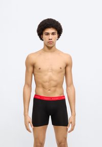 Μαύρα ανδρικά μποξέρ με κόκκινη μέση που φέρει το λογότυπο "TOMMY HILFIGER", με λεία υφή και εφαρμοστό σχεδιασμό, παρουσιασμένα σε αρσενικό μοντέλο.