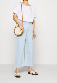 Kevyt valkoinen pellavaposliinipaita, jossa on lyhyet hihat, yhdistettynä korkeavyötäröisiin, leveisiin vaaleansiinisiin farkkuihin. Ruskea olkalaukku ja sandaalit.