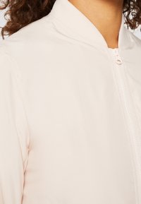 Gros plan d'une personne portant une veste zippée beige clair avec un col côtelé et des cheveux bouclés visibles sur le bord.