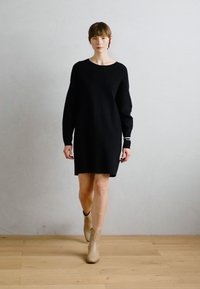 Vestido de punto negro con manga larga, escote redondo y un corte relajado, combinado con botas de tobillo beige sobre un suelo de madera.