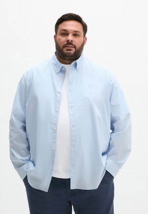 Mann mit Bart, der ein hellblaues Hemd über einem weißen T-Shirt und dunklen Hosen trägt, Hände in den Taschen, neutraler Hintergrund.