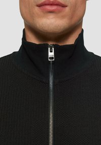 Schwarze Zip-Jacke mit hohem Kragen, strukturiertem Stoff und silbernem Reißverschluss, der ein Logo am Zipper aufweist.