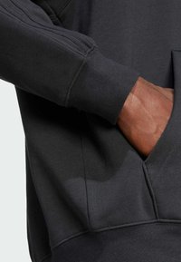 Black sweatshirt fabric with a textured surface, featuring a kangaroo pocket and ribbed cuffs. Close-up on sleeve and hand placement.