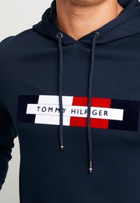 Granatowy hoodie z miękkiej bawełny z naszywką na przodzie, na której widoczne są czerwone, białe i granatowe paski. Regulowany sznurkiem z metalowymi końcówkami.
