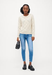 Kreminė megzta suknelė su deimanto raštais ir nedideliais akcentais, derinama su šviesiai mėlynomis liesomis kelnėmis ir juodomis platforminėmis bateliais. Juodas rankinukas laikomas rankoje.