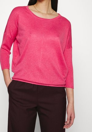 Kobieta w luźnym różowym swetrze z rękawami długości trzy czwarte zakończonymi ściągaczami i ciemnobrązowych spodniach, z rękami w kieszeniach, stojąca na białym tle.