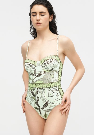 Jeune femme aux longs cheveux foncés portant un maillot de bain une pièce vert pâle à motifs botaniques et d'oiseaux.