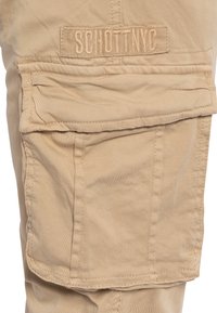 Beżowe spodnie cargo z dużą boczną kieszenią na klapę. Tkanina jest gładka z subtelnym połyskiem i zawiera teksturowaną metkę z napisem "SCHOTT NYC".