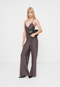 Salopetă strălucitoare de culoare mov închis, cu un corset ajustat, picioare larghi de palazzo și bretele subțiri. Este asortată cu o poșetă neagră și tocuri.