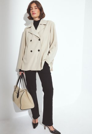 Beige dubbelknäppt jacka med svarta knappar, svart polotröja, svarta byxor och eleganta svarta klackar. Håller en beige tote-väska med svarta accenter.