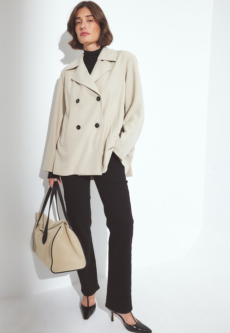 Beige double-breasted jacket with black buttons, black turtleneck, black pants, and sleek black heels. Holding a beige tote bag with black accents.