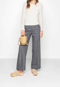 Beżowy sweter dziany z falbankowym dekoltem, szare spodnie o szerokich nogawkach i beżowe chodaki. Trzyma małą, okrągłą torebkę w kolorze beżowym z kolorowym akcentem.