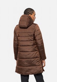 Vrouw met gevlochten haar in een bruin gewatteerd hooded coat, staande terzijde tegen een effen achtergrond.