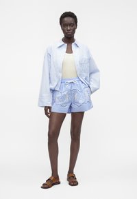 Femme portant une chemise oversize rayée bleu clair, un débardeur crème, un short bleu brodé et des sandales marron, debout devant un fond blanc.