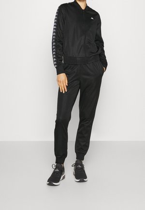 Person wearing black zip-up tracksuit with logo stripes on sleeves and black sneakers, standing with hands in pockets.
