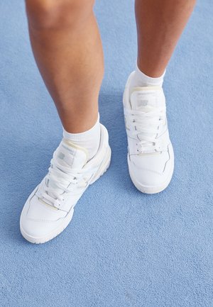 Person trägt weiße New Balance Sneakers und weiße Socken und steht auf einem hellblauen Teppichboden.