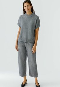 Eine Frau trägt ein hellgraues kurzärmliges Strickoberteil und weit geschnittene graue Jeans mit verkürzter Beinlänge. Sie steht mit entspannter Haltung vor einem weißen Hintergrund.