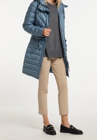 Jasnoniebieska kurtka puchowa z kapturem, pikowany wzór i ściągacz w talii. Założona na szary sweter i beżowe spodnie, zestawiona z czarnymi mokasynami.