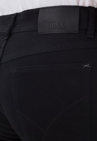 Gros plan de la poche arrière d'un pantalon noir avec des passants pour ceinture et un patch en cuir de la marque BRAX au-dessus de la poche sur un fond en tissu blanc.