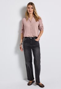 Frau, die gegen eine weiße Wand steht, trägt ein hellrosa Hemd mit Knöpfen, schwarze hochtaillierte Jeans, silberne Armbänder und schwarze Netz-Ballerinas.