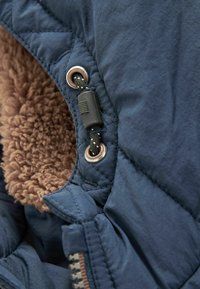 Marineblaue isolierte Jacke aus Stoff, mit einem weichen braunen Kunstfellfutter, gestepptem Muster und schwarzem Kordelzug mit silbernen Ösen.