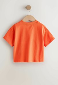 Oranžové tričko z bavlny s krátkym rukávom a okrúhlym výstrihom, zavesené na drevenom ramienku na svetlom pozadí. Bez ozdôb alebo vzorov.