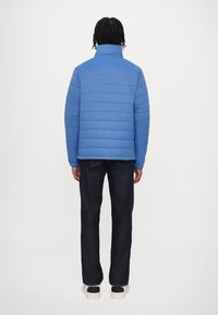 Blue puffer jacket with a high collar, featuring horizontal quilting and long sleeves. Paired with dark denim pants and white-soled shoes.
