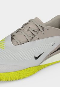 Μπεζ και λευκό αθλητικό παπούτσι Nike με υφασμάτινο επάνω μέρος, μαύρο λογότυπο Swoosh, κείμενο "NIKESKIN", γκρι κορδόνια και φωτεινή κίτρινη ελαστική σόλα.