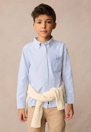 Camicia a righe con bottoni in azzurro chiaro e bianco, con un maglione crema a trecce annodato in vita, abbinata a pantaloni beige.