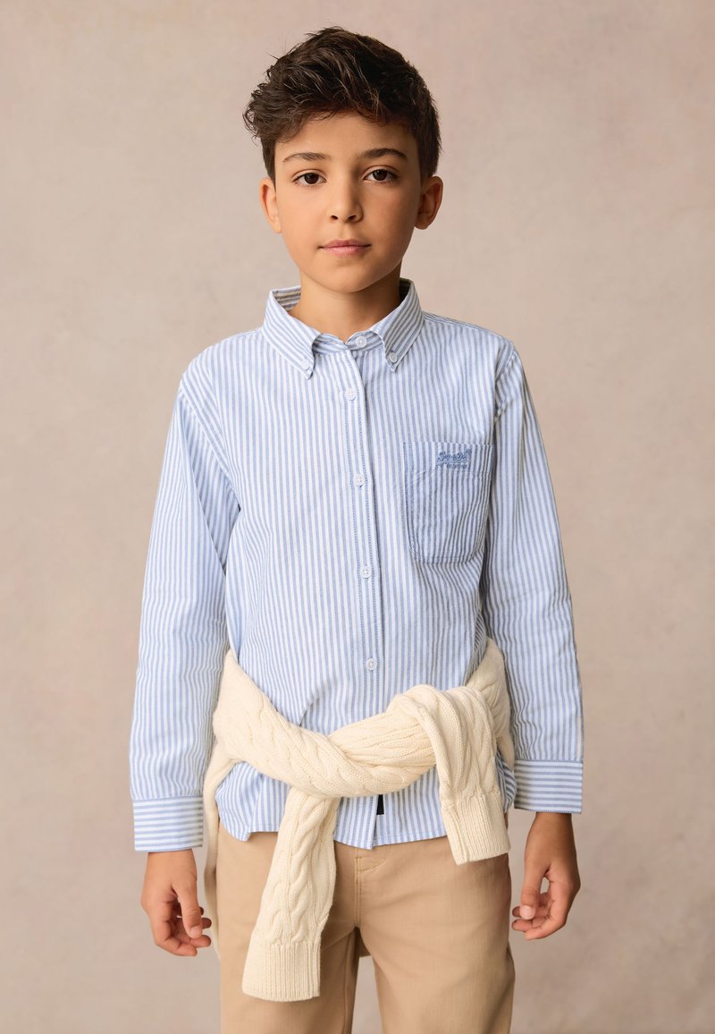 Világoskék és fehér csíkos ing, ráhúzott krémszínű fonott mintás pulóverrel a derékon, bézs nadrággal párosítva.