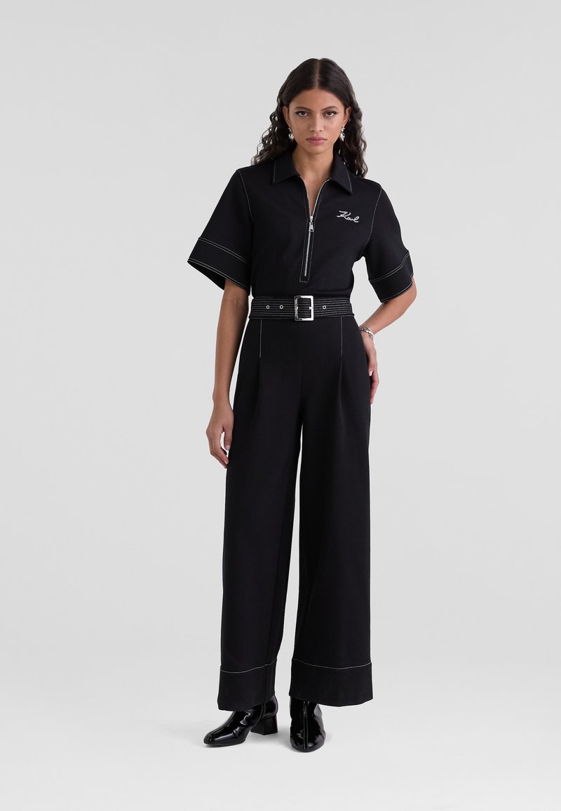 Salopette noire à manches courtes, col zippé, couture blanche, jambes larges et ceinture assortie. Portée avec des bottes noires à talons.