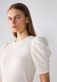 Top alb texturat cu mâneci umflate. Țesătura este moale, cu un model subtil și un decolteu rotund.