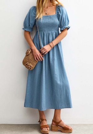 Kvinde iført en blå midi-kjole med pufærmer, brune platformsandaler og en vævet crossbody-taske, stående foran en hvid væg.