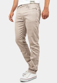 Pantalon beige à rayures en tissu léger, coupe slim, avec poches avant, fermeture à bouton, associé à des baskets blanches.