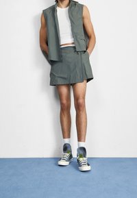Gilet grigio senza maniche sopra una canotta bianca, abbinato a una gonna grigia coordinata. Sneakers grigie con dettagli verdi e calze a fantasia.