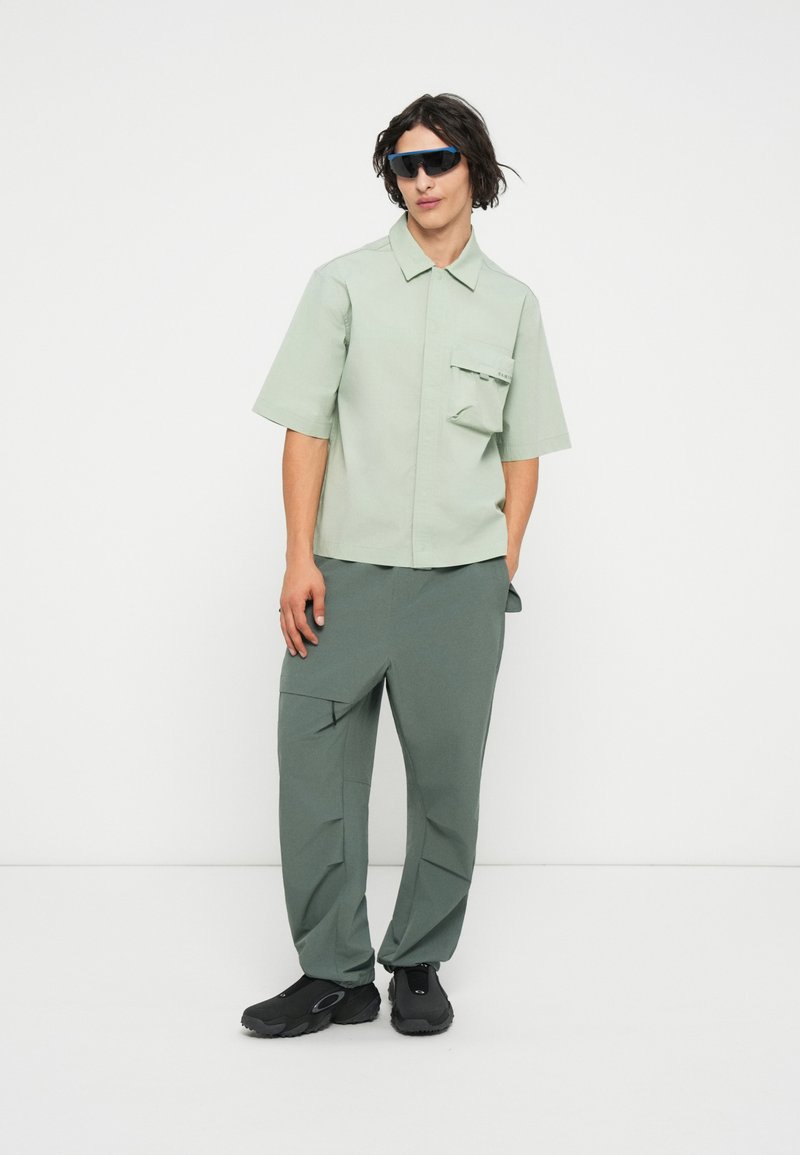 Camisa de cuello corto de manga corta verde claro con un bolsillo en el pecho; combinada con pantalones cargo holgados verde oscuro y zapatillas deportivas negras.