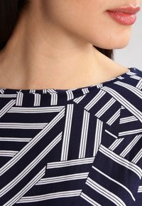 Marinblått tyg med vita randiga geometriska mönster, slät textur och vid halsringning på ett plagg. Närbild av design detaljer.